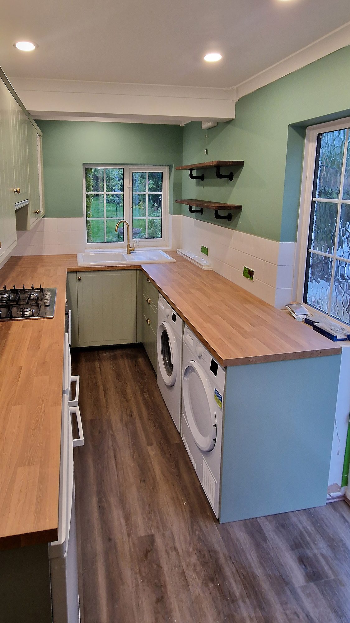 U-shaped kitchen in Worcester Park with sage green units, oak-effect worktop, sink under the window and washing machine beside a tumble dryer