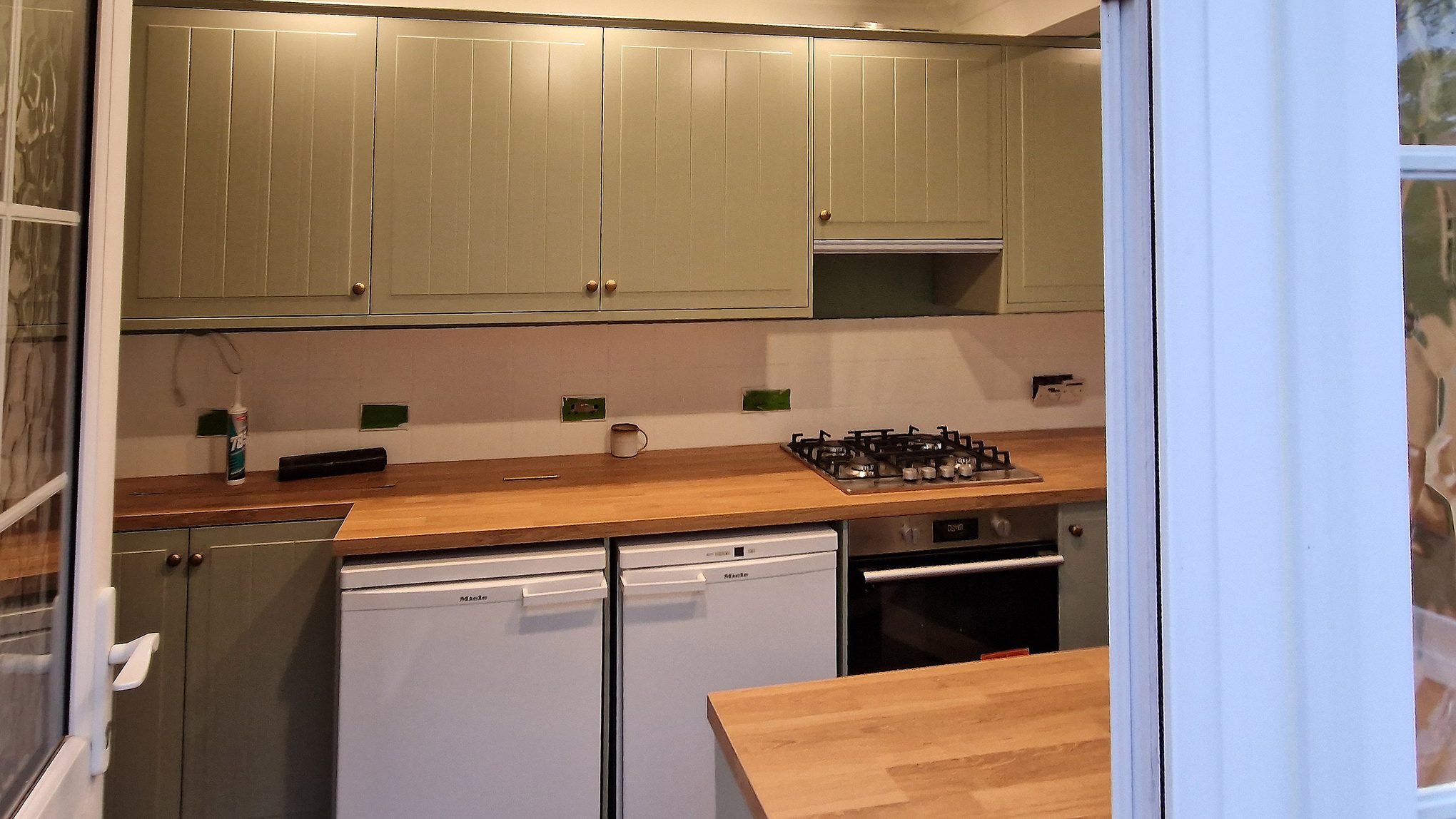 Close view of sage green panelled wall units, brass knobs, gas hob and under-counter appliances in a Worcester Park kitchen