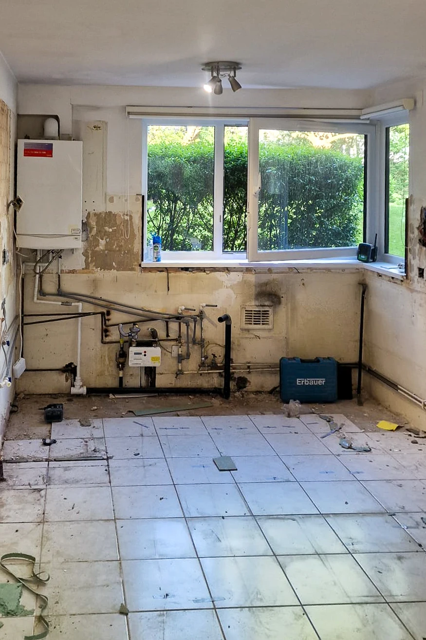 Kitchen in Wandsworth fully stripped before renovation, showing exposed gas pipes, boiler and bare plaster walls